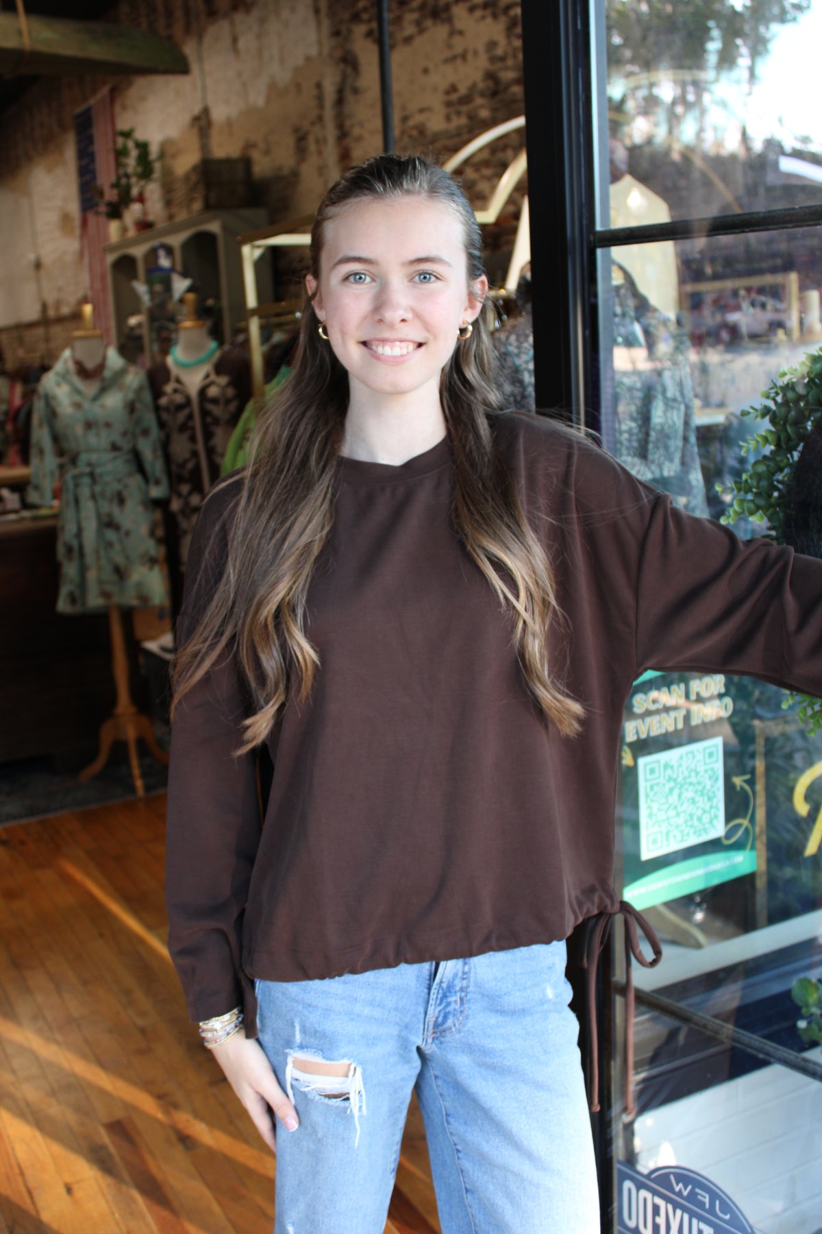 A young woman with long brown hair stands in a clothing boutique wearing a dark brown long-sleeve top and light blue distressed jeans.