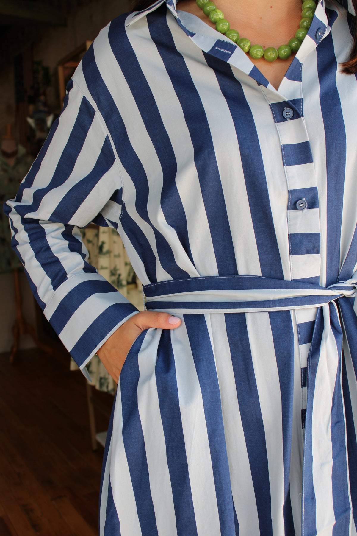 A close-up of a person wearing a blue and light blue vertically striped long-sleeve shirt dress with a matching tie belt, paired with a green beaded necklace.
