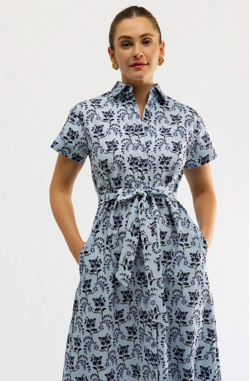 A woman modeling a light blue short-sleeve collared dress with a dark navy floral print and a matching waist tie, set against a plain light gray studio background.