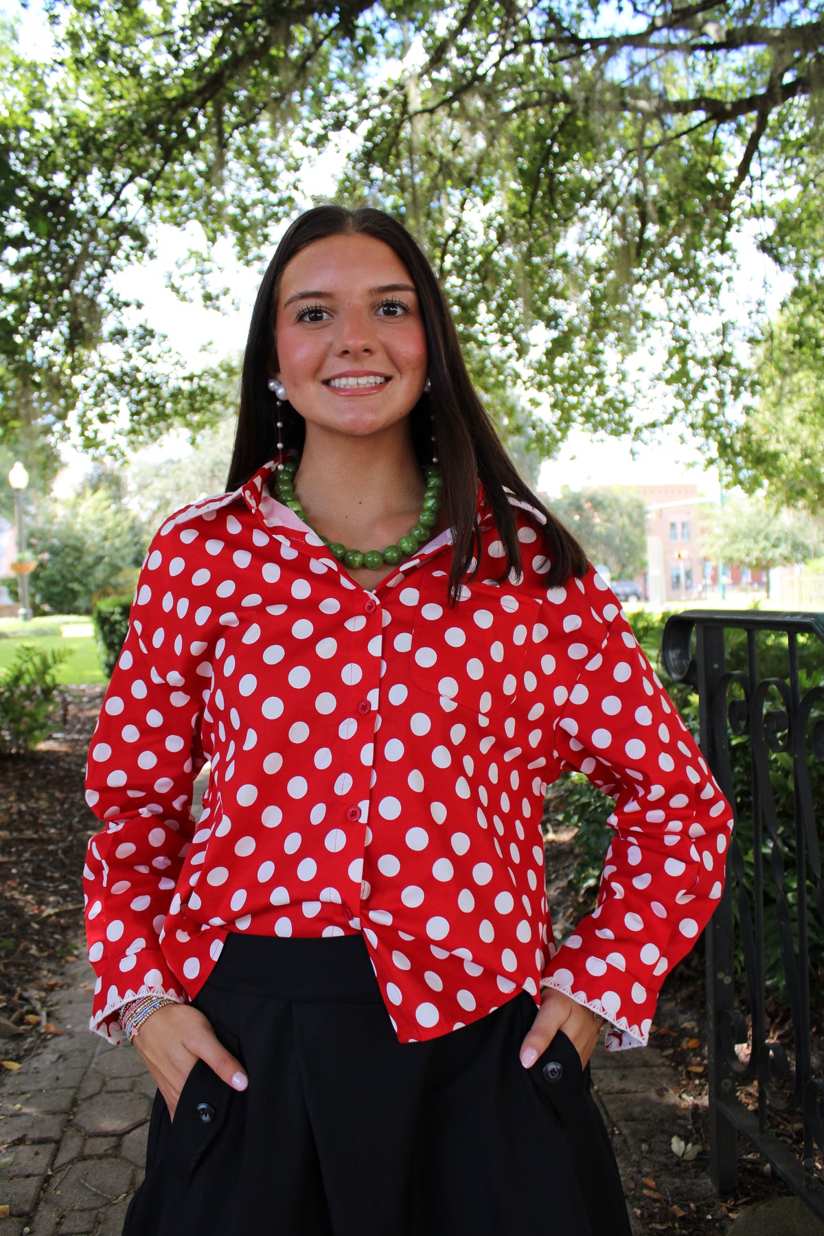 A young woman smiling outdoors under a tree, wearing a red long-sleeve button-up blouse with white polka dots and black pants.
