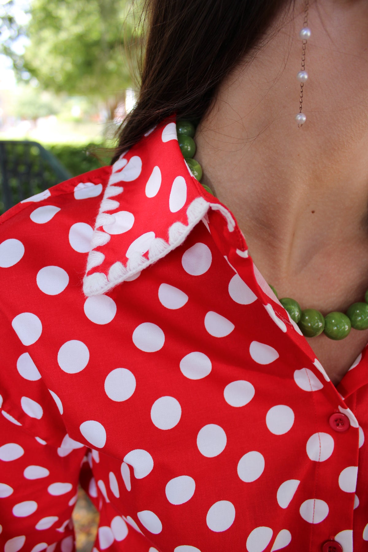 Close-up of a person wearing a red collared button-up shirt with large white polka dots, along with a green beaded necklace and dangling earrings, outdoors with blurred greenery in the background.