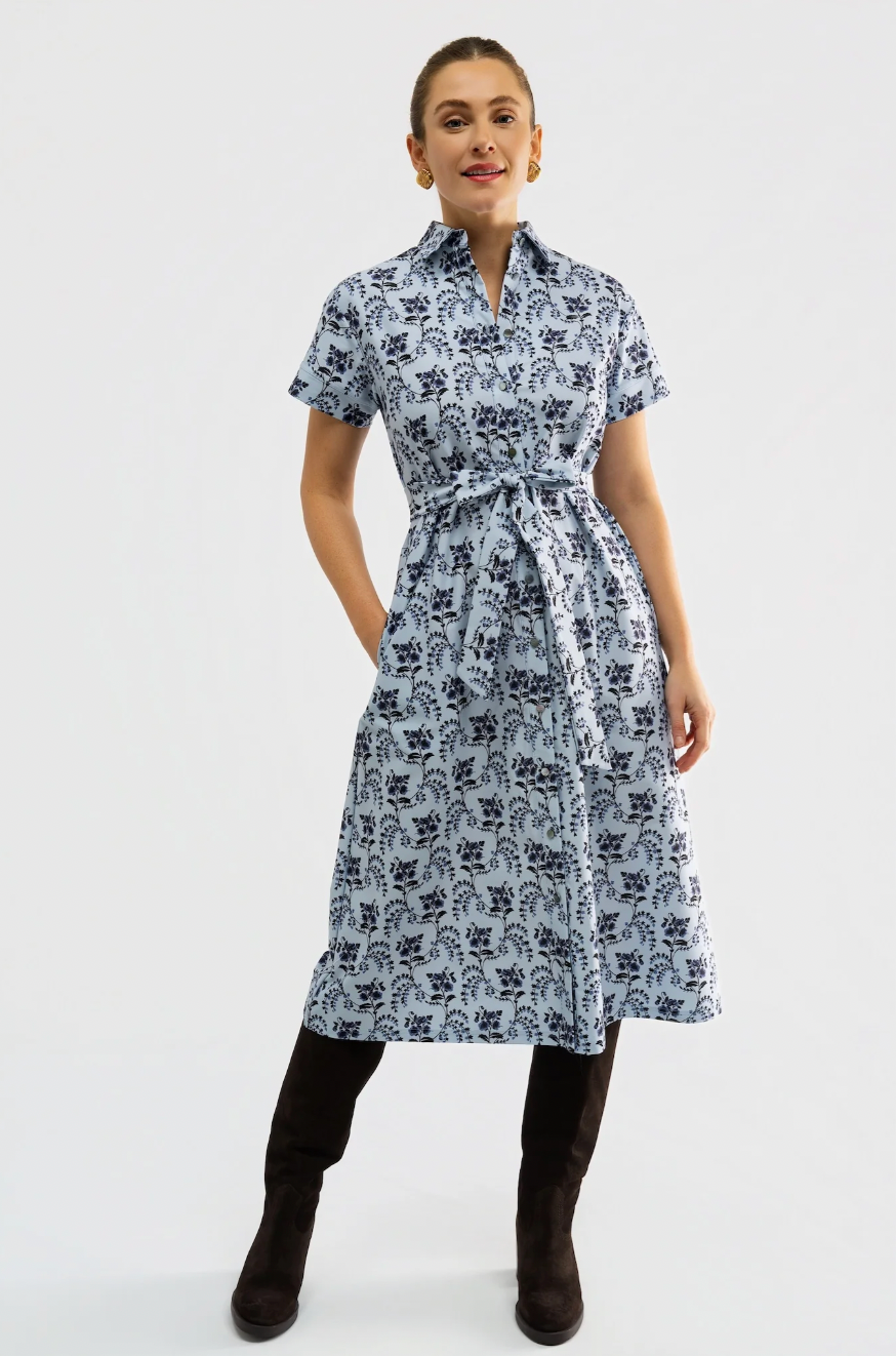 A woman modeling a short-sleeve midi dress with a dark blue floral block print on a light blue-gray background, featuring a matching waist tie belt. She is wearing dark brown knee-high boots against a plain white studio background.