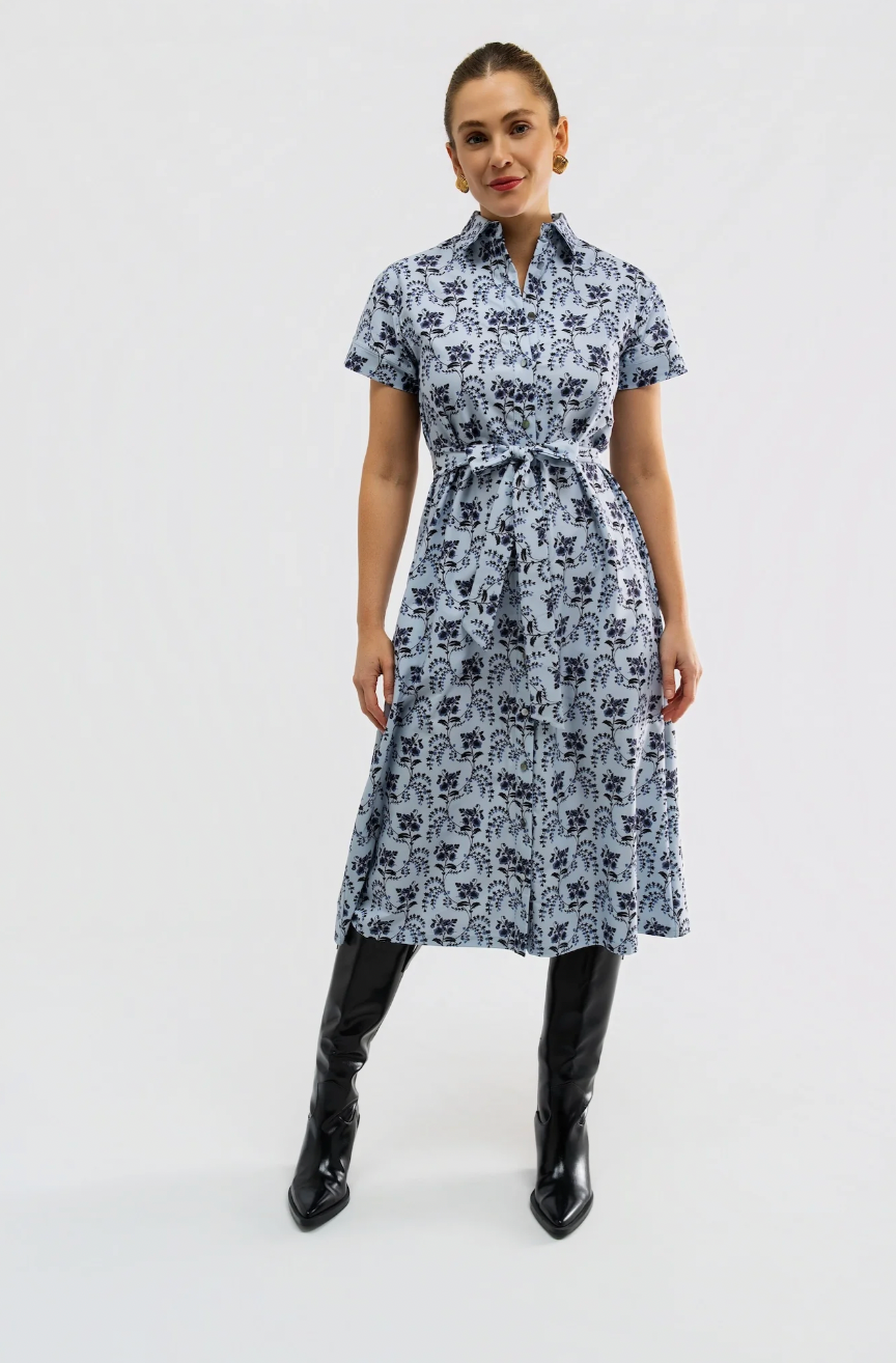 A woman modeling a short-sleeve midi dress with a dark blue floral block print on a light blue-gray background, featuring a matching waist tie belt. She is wearing dark brown knee-high boots against a plain white studio background.