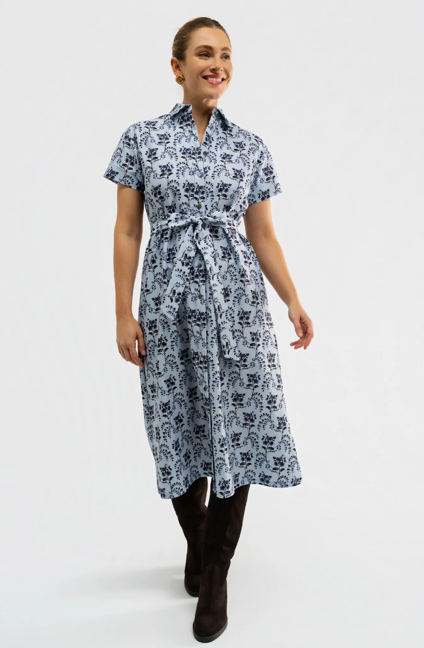 A woman modeling a short-sleeve midi dress with a dark blue floral block print on a light blue-gray background, featuring a matching waist tie belt. She is wearing dark brown knee-high boots against a plain white studio background.