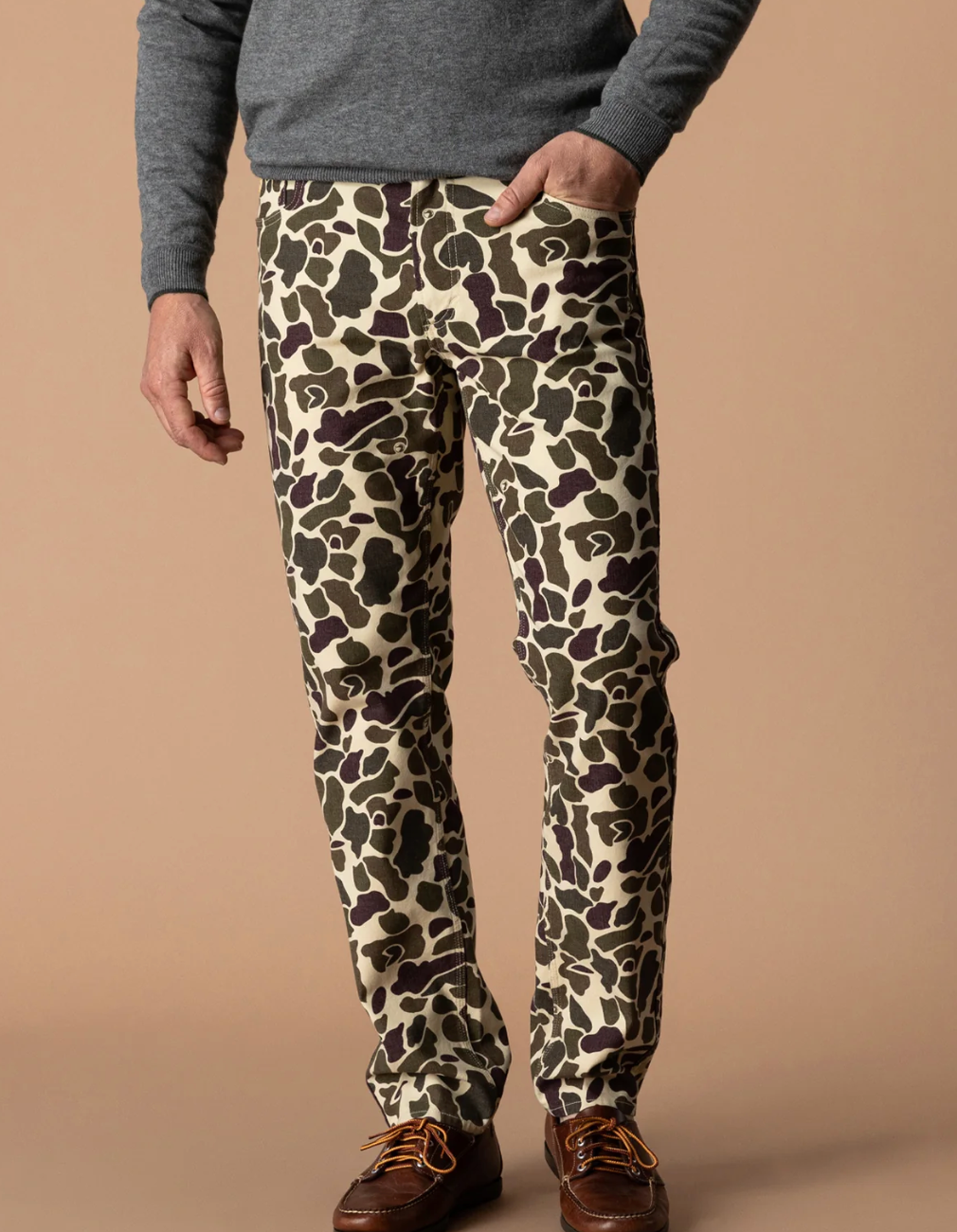 A person wearing slim-fit trousers with an all-over camouflage pattern in beige, olive green, and dark brown, styled with a gray sweater and brown leather shoes against a solid beige studio background.