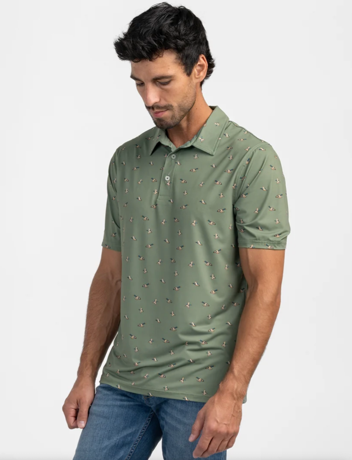 A man wearing a sage green short-sleeve polo shirt with a small all-over printed pattern of mallards, paired with blue jeans, photographed against a plain light gray studio background.