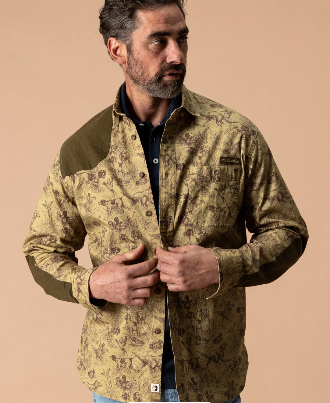 A man wearing a long-sleeve button-up shirt jacket with a brown and beige deer hunting pattern, brown shoulder panels, and a dark navy polo shirt underneath, set against a solid beige background.