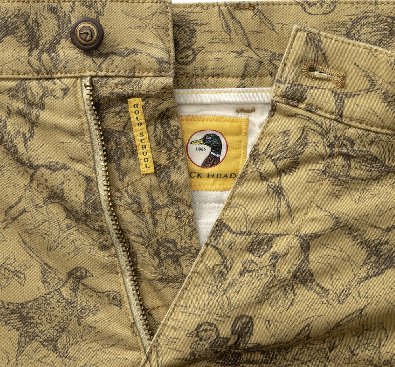 Close-up of a tan and brown duck hunting patterned jacket with a zipper, button, and a yellow neck label featuring a duck head logo and the text 'Duck Head'.