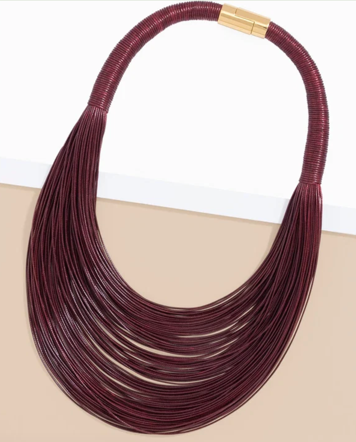 A multi-strand statement necklace made of dark burgundy cord with a gold clasp, displayed on a split white and beige background.