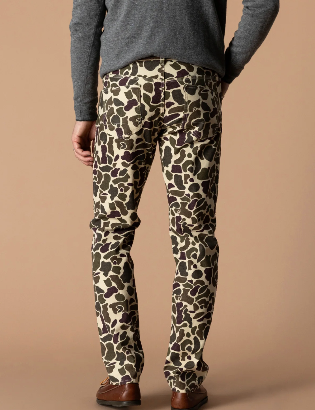 A person shown from the back wearing slim-fit trousers with an abstract organic spot pattern in beige, olive green, and dark brown, paired with a gray sweater and brown shoes against a plain beige studio background.