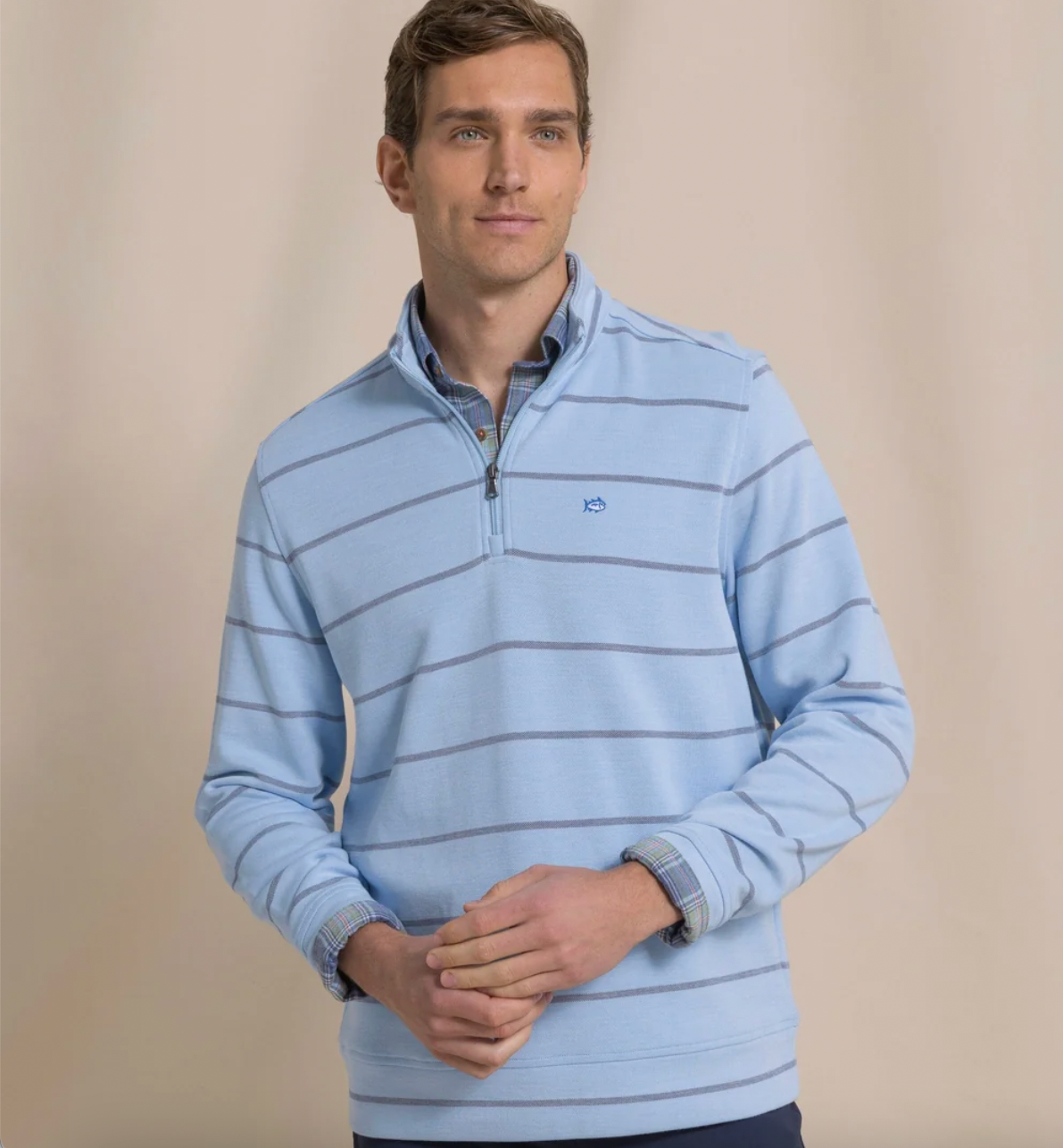A man wearing a light blue quarter-zip pullover sweater with thin dark horizontal stripes, standing against a neutral beige studio background.