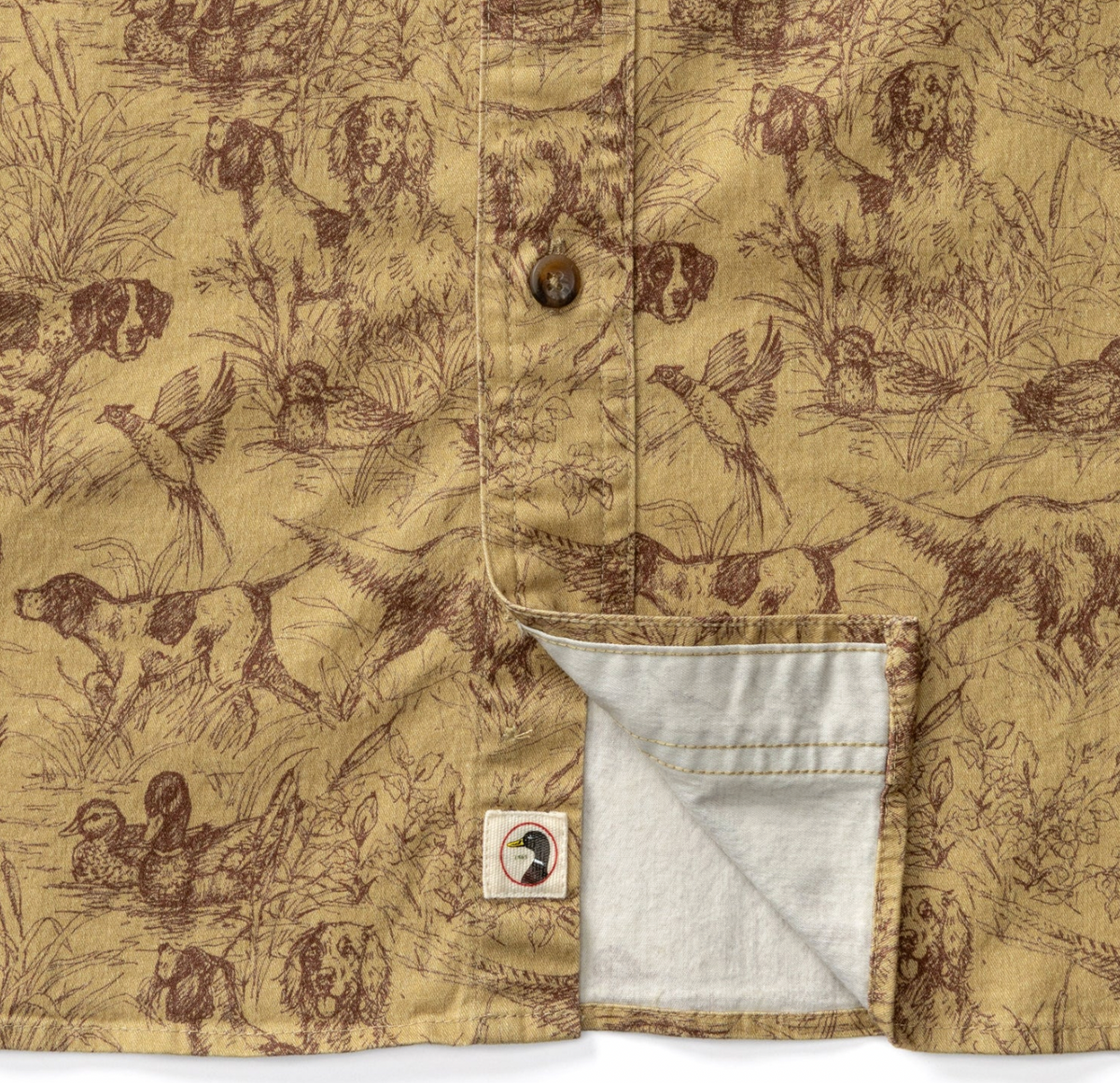 Close-up of a button-front shirt with a tan and brown all-over hunting-themed print featuring dogs, ducks, birds, and reeds. A pocket is shown open to reveal a light gray lining, and a small square brand patch is visible near the pocket.