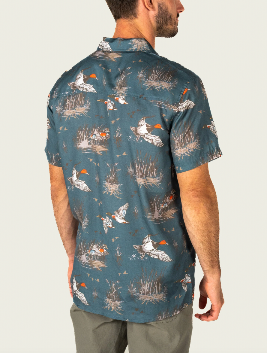 A man wearing a short-sleeve button-up shirt with a detailed duck hunting print, shown from the back against a light gray studio background.
