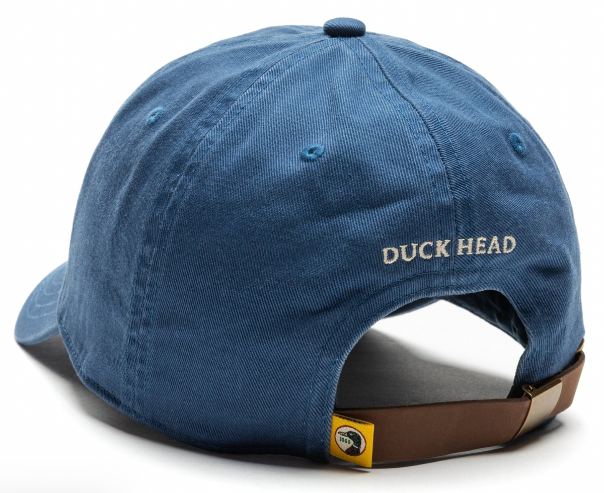 A blue cotton baseball cap shown from a three-quarters back angle, featuring 'DUCK HEAD' embroidered in beige on the side and a brown adjustable strap with a small yellow Duck Head logo tag.