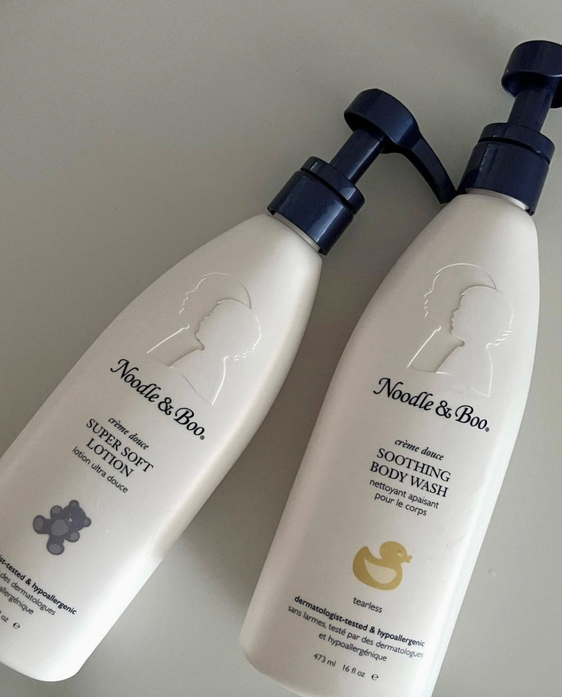 Two white pump bottles of Noodle & Boo baby care products on a light gray background: Super Soft Lotion and Soothing Body Wash.
