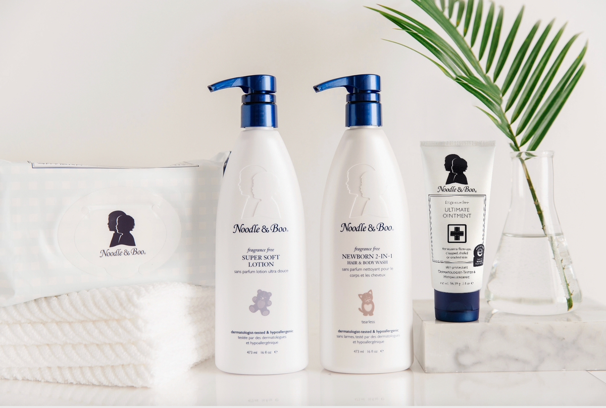 Three Noodle & Boo baby care products arranged on a white surface with a folded white towel, a green palm leaf, and a clear glass vase. The set includes a pump bottle of Super Soft Lotion, a pump bottle of Baby Body Wash, and a tube of Ultimate Ointment.