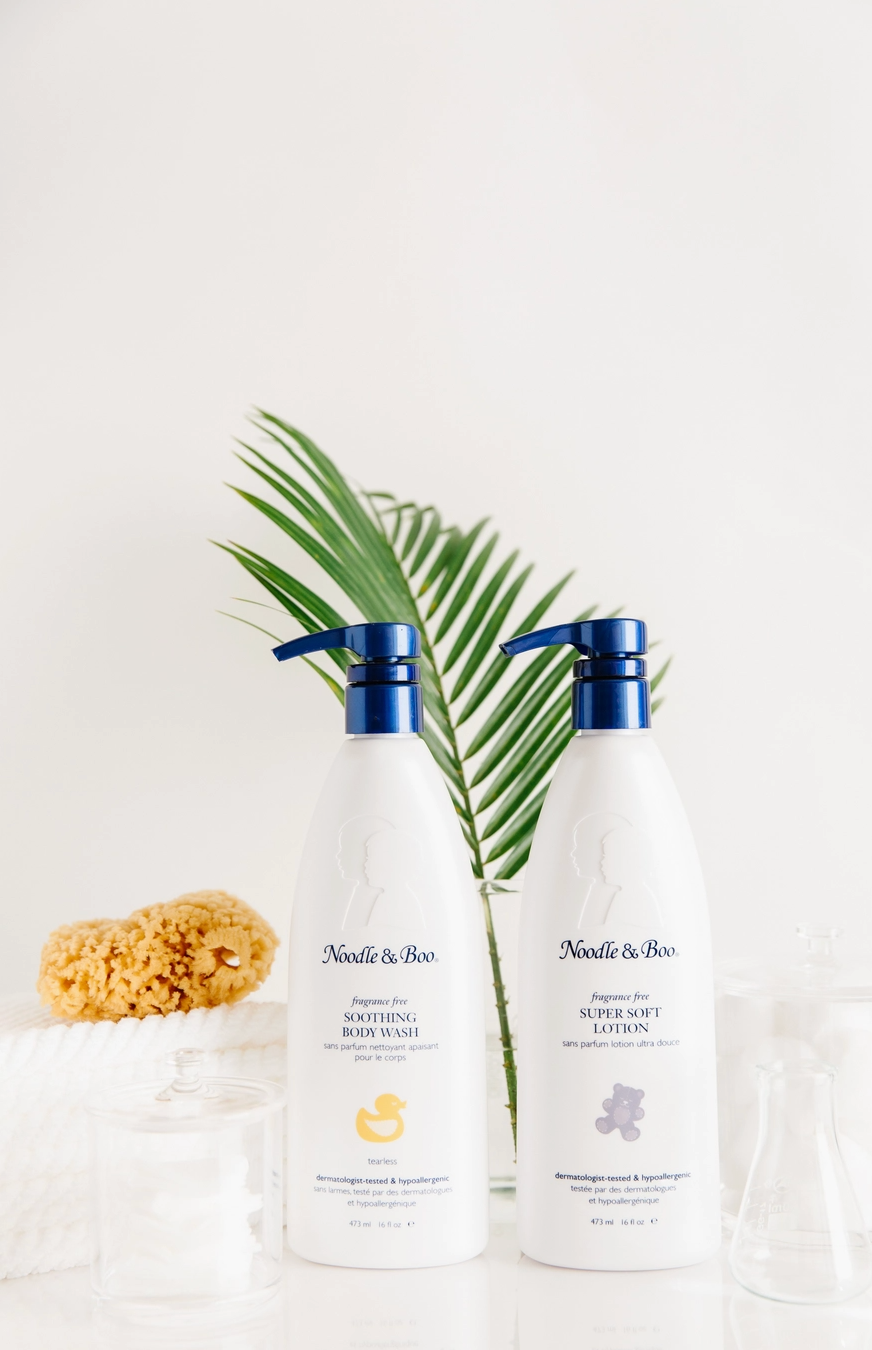 Two white bottles of Noodle & Boo body care products with blue pumps, styled with a green palm leaf and a sponge on a white background.