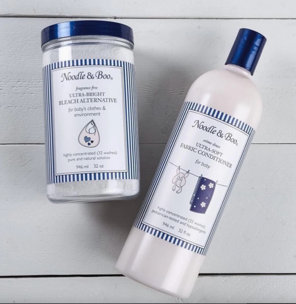 Two Needle & Boo baby care products on a white wood background: a clear plastic jar of ultra-bright bleach alternative with a blue lid and label, and a white bottle of fabric conditioner with a blue cap and label.
