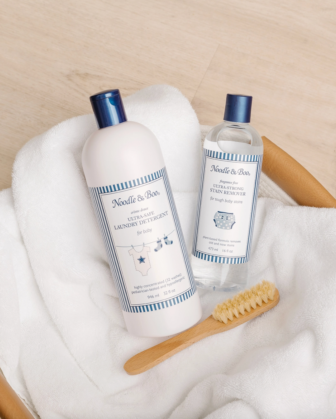 Two Needle & Rose laundry care products arranged on white towels with a wooden brush on a light wood surface. The set includes a large bottle of ultra soft laundry detergent and a smaller bottle of softening foam, both with white labels and blue caps.