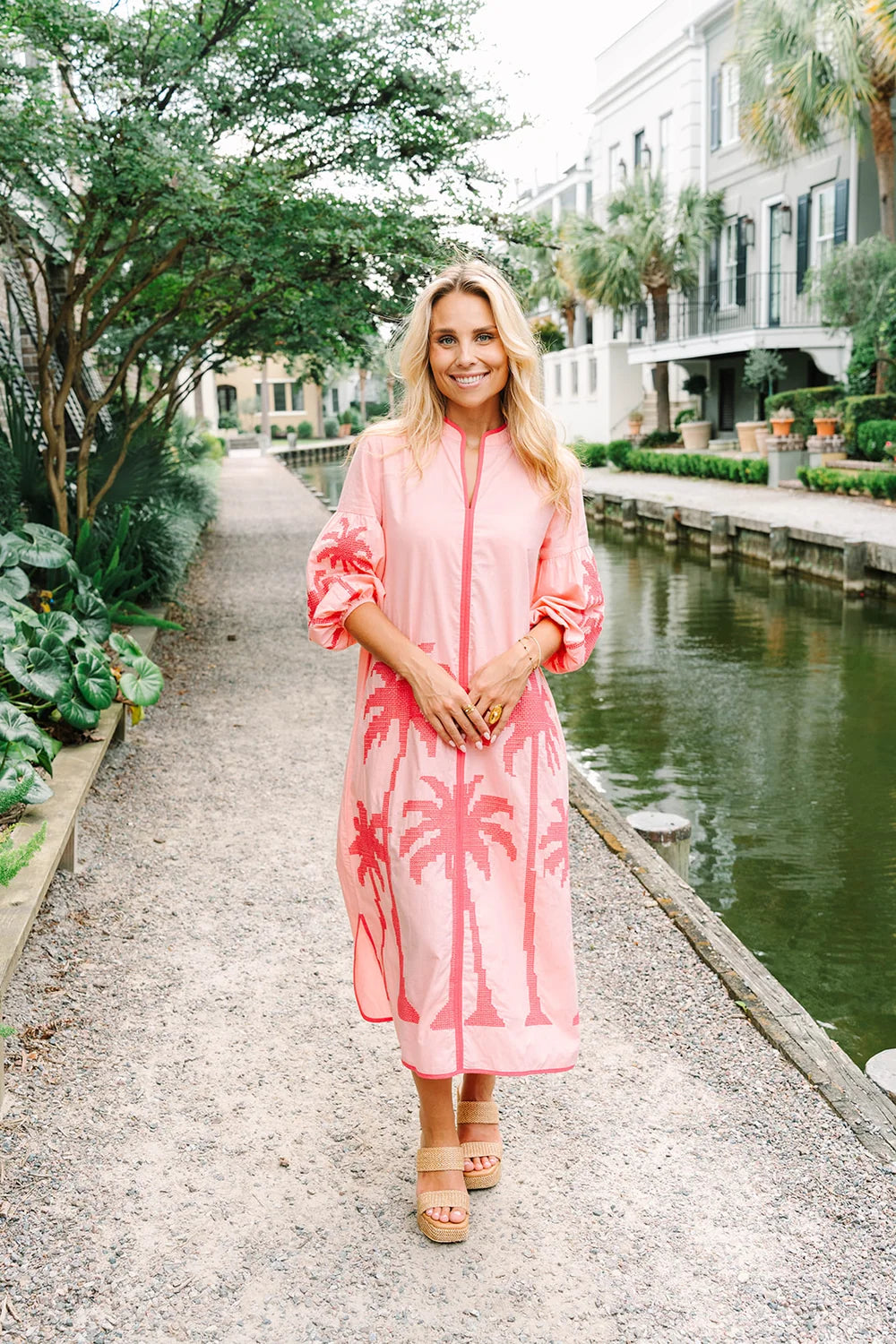 Woman in a pink dress standing by a canal with buildings and greenery in the background