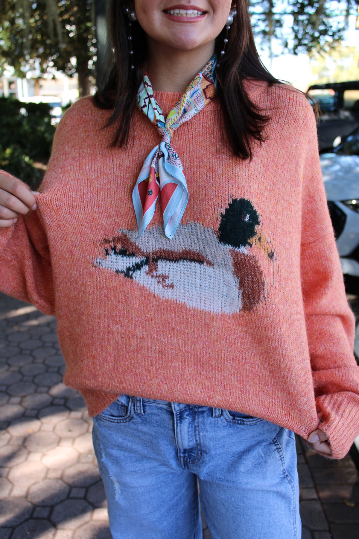 A woman wearing a fuzzy coral-pink knit sweater with a large duck motif across the front, styled with a patterned neck scarf and blue jeans in an outdoor setting.