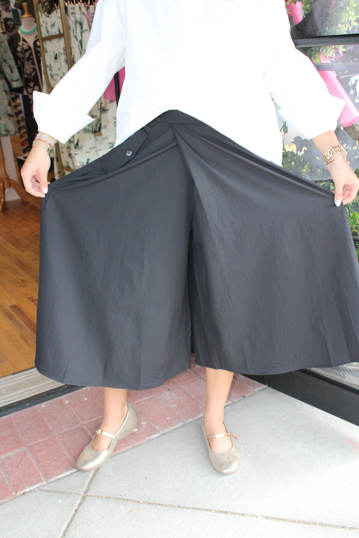A person wearing black wide-leg cropped pants or culottes, holding them out to show the full shape. The person is also wearing a white long-sleeve top and metallic flat shoes, standing on a tiled floor in front of a shop window.