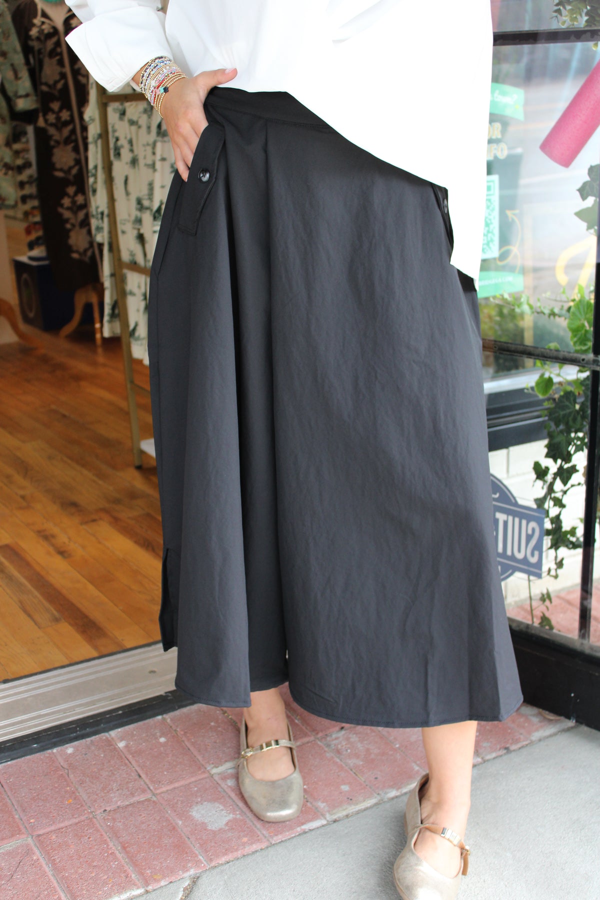 A person wearing a blackwide-leg midi pant that gives the illusion of a skirt with a white top and metallic flat shoes, standing in front of a boutique window.