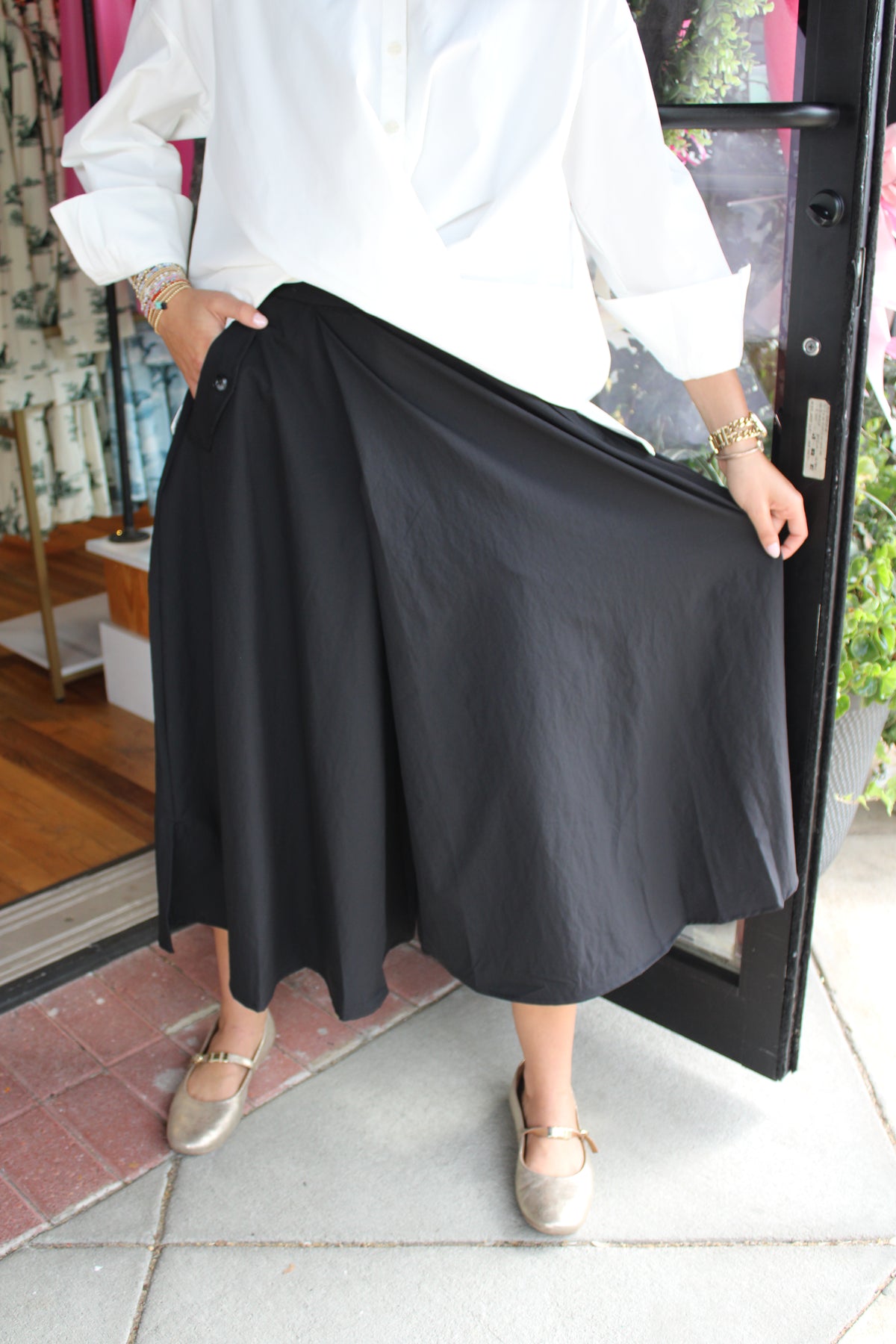 A person wearing a loose white long-sleeve top and a black midi pant, holding out the skirt to show its drape. The person is standing on a tiled sidewalk in front of a storefront.