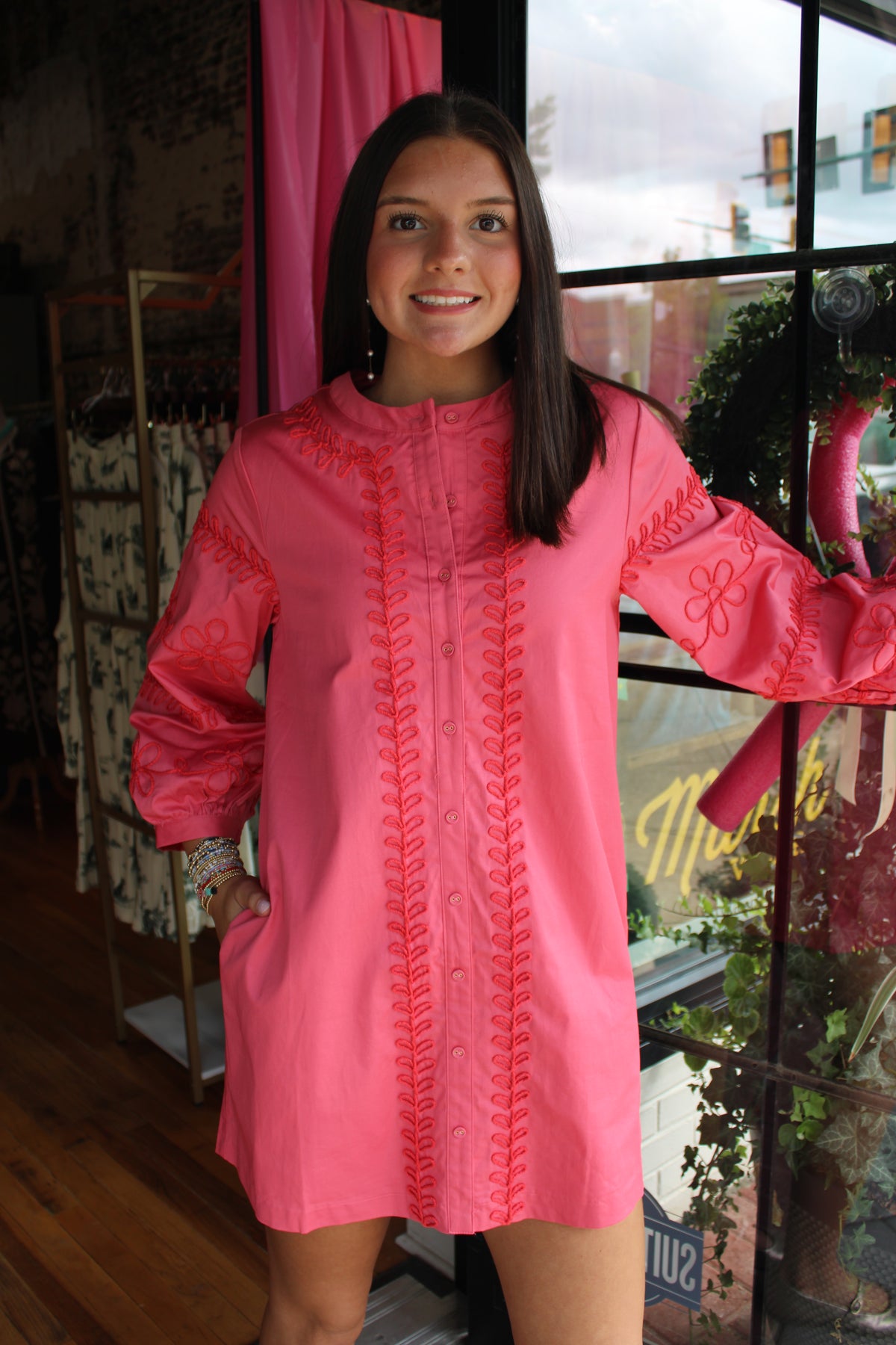 A woman modeling a bright pink long-sleeve tunic dress with matching pink embroidery, standing in a boutique setting.