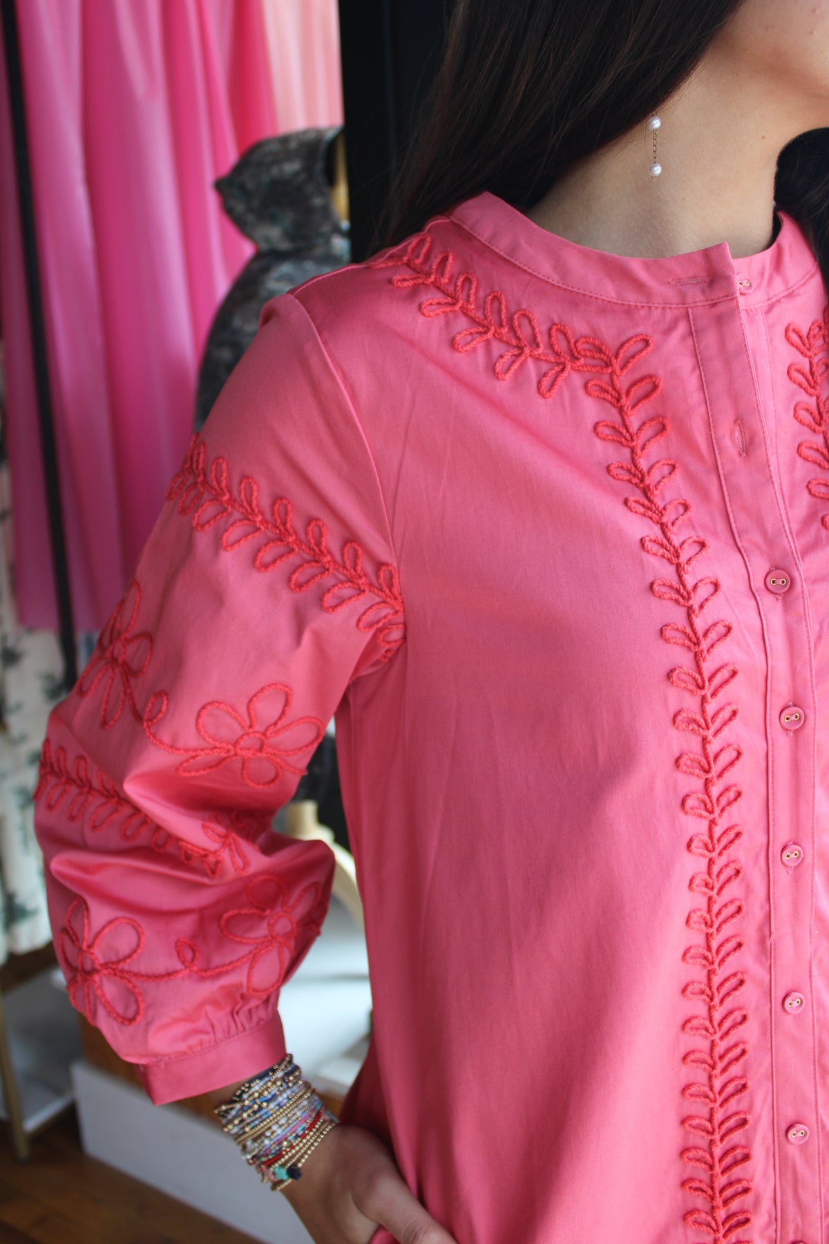 A close-up of a woman wearing a bright pink long-sleeve blouse with red embroidered floral and leaf patterns along the front and sleeves.