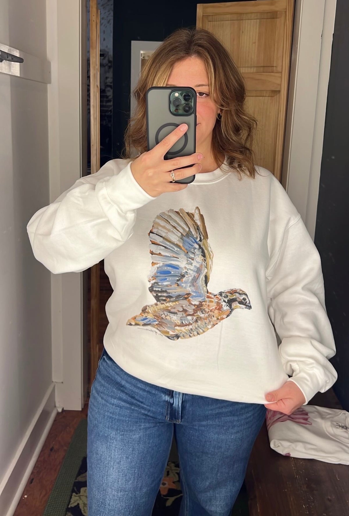 A woman takes a mirror selfie wearing a white crewneck sweatshirt with a large, detailed graphic of a flying quail in shades of blue, brown, and gold, paired with blue jeans.