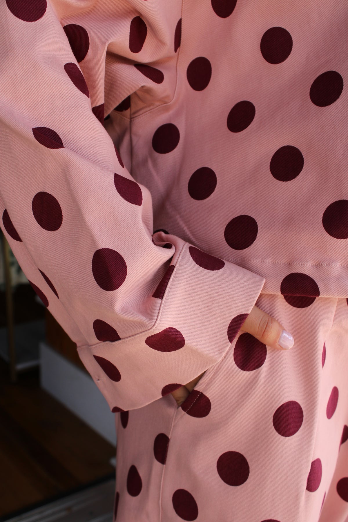 A close-up of a person wearing a pink long-sleeve jacket with large dark burgundy polka dots, with one hand tucked into a pocket.