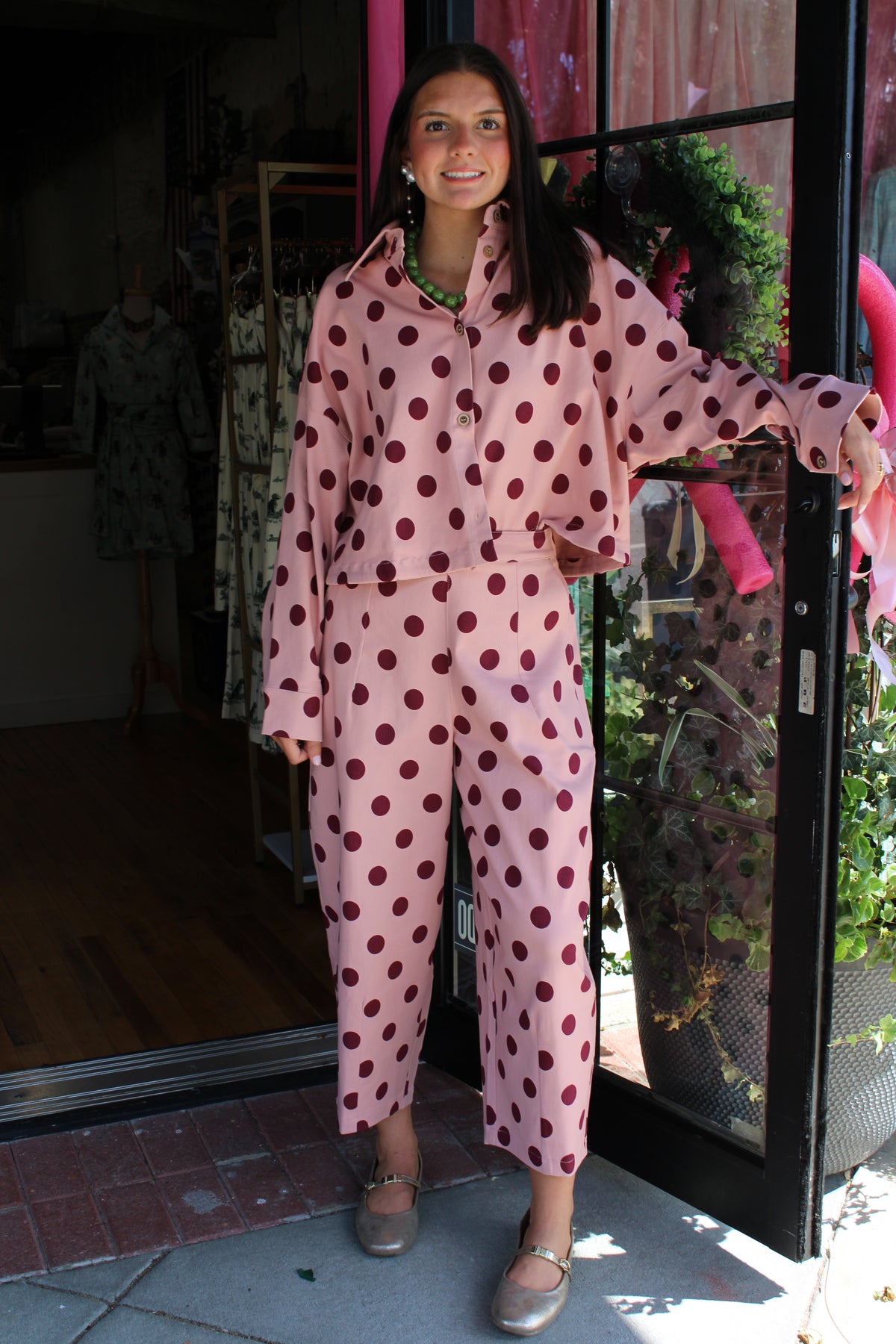 A woman stands in a storefront doorway wearing a matching pink set with dark red polka dots. The set includes a long-sleeve button-front jacket and pants.