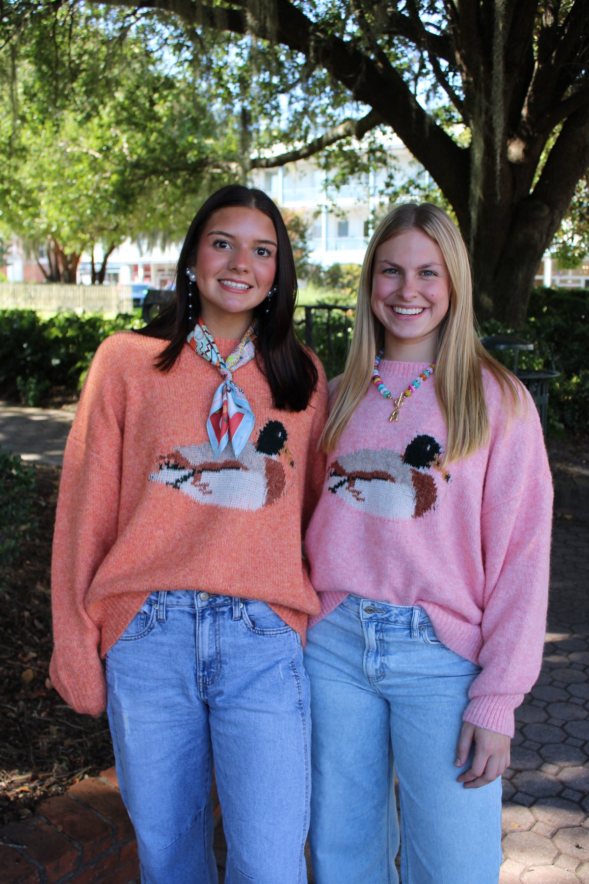 Two women standing outdoors in front of trees, wearing matching knit sweaters with a duck design. The woman on the left wears a cider sweater and a patterned neck scarf, while the woman on the right wears a light pink sweater.