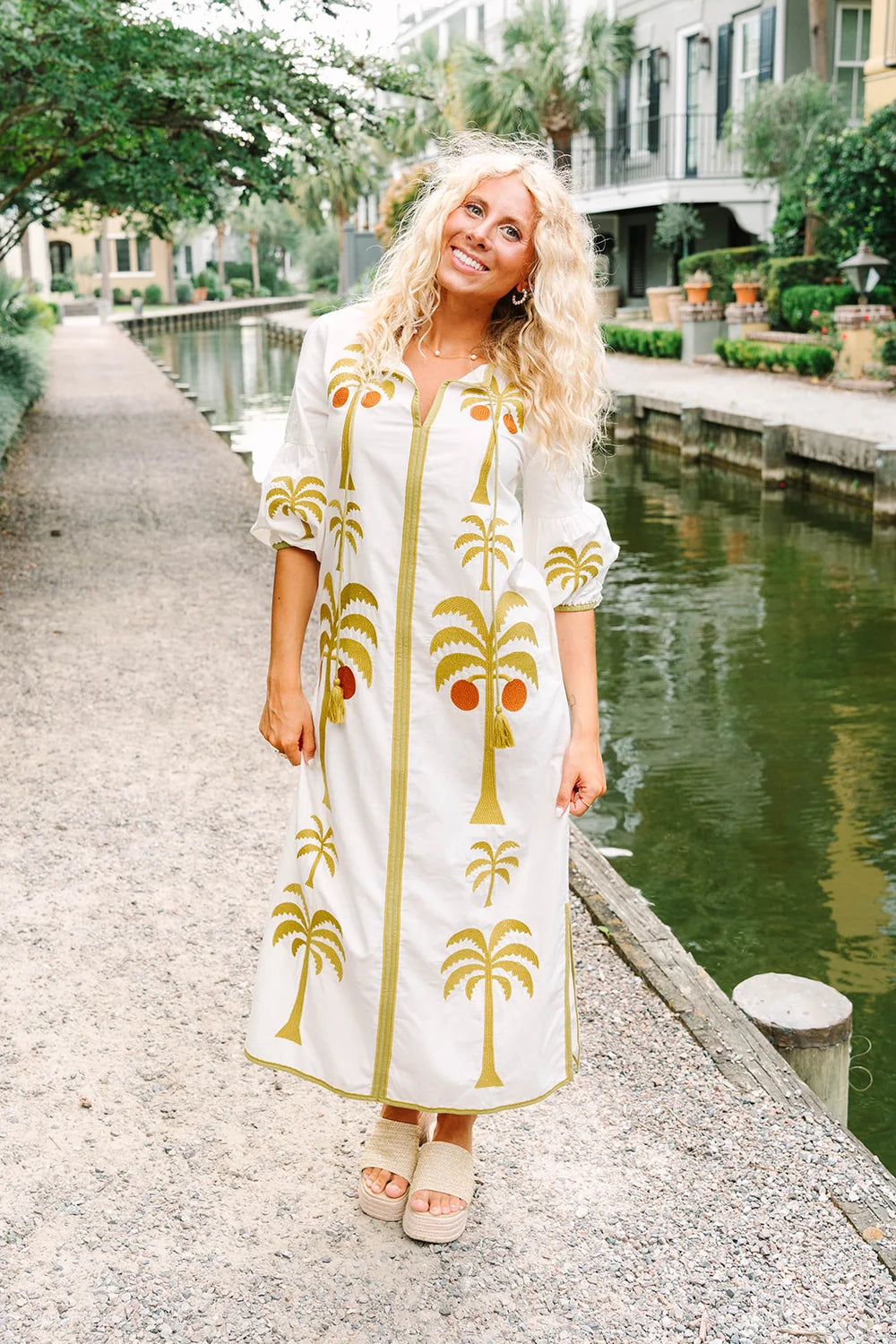 Woman in a white dress with palm tree patterns standing by a canal.