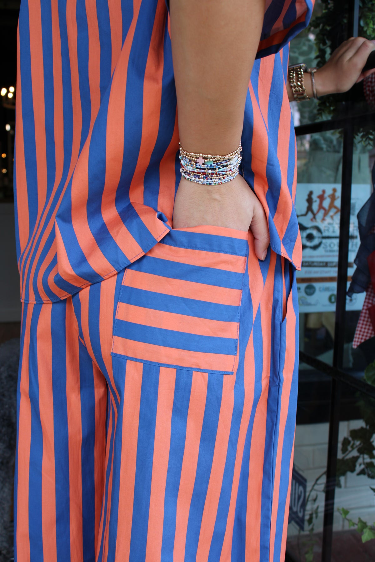 A close-up of a person wearing a loose-fitting striped pant with vertical blue and orange stripes, featuring a large patch pocket on the thigh and a side pocket. 