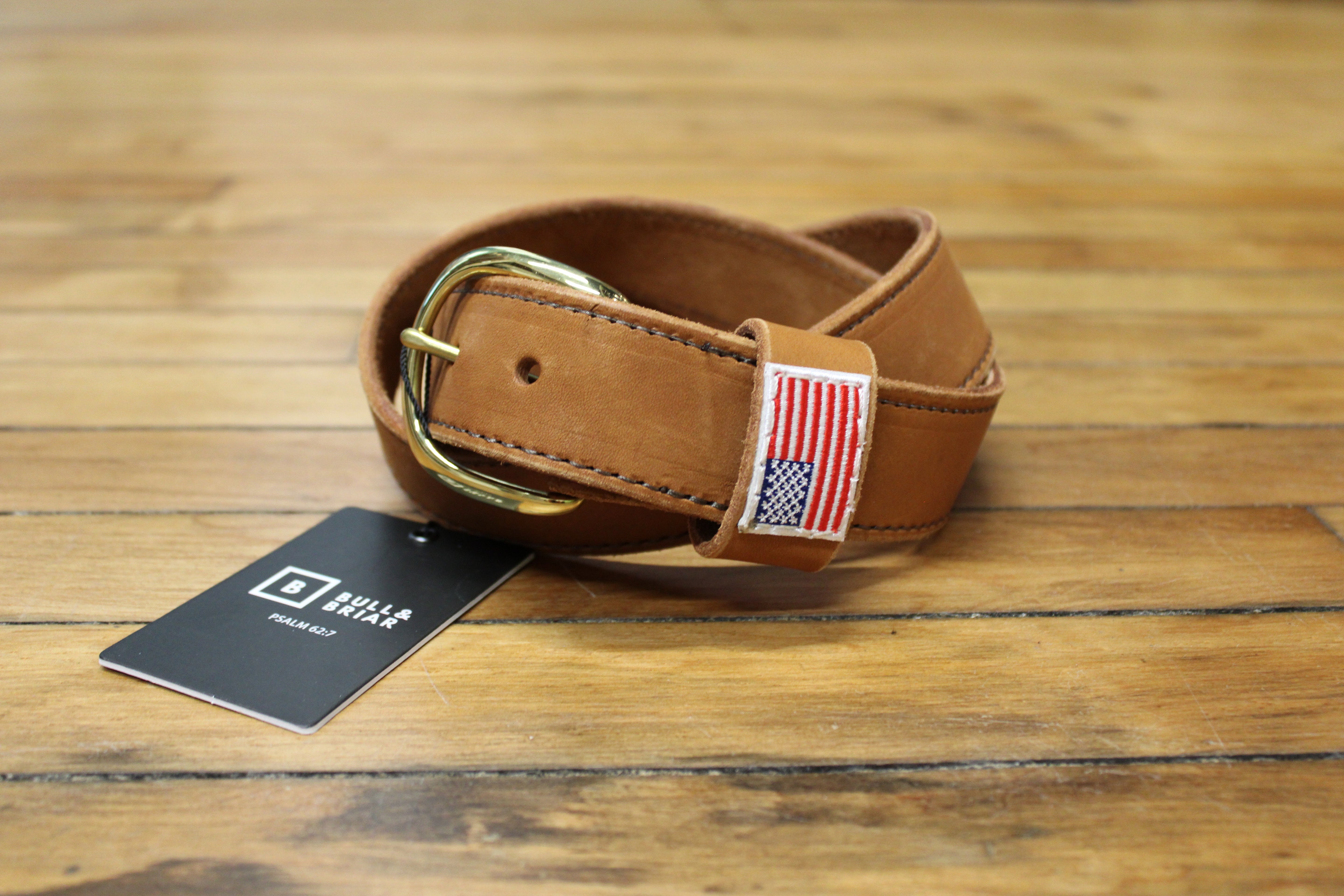 A tan leather belt with a gold buckle, featuring a small American flag patch on the buckle loop and a black brand hangtag