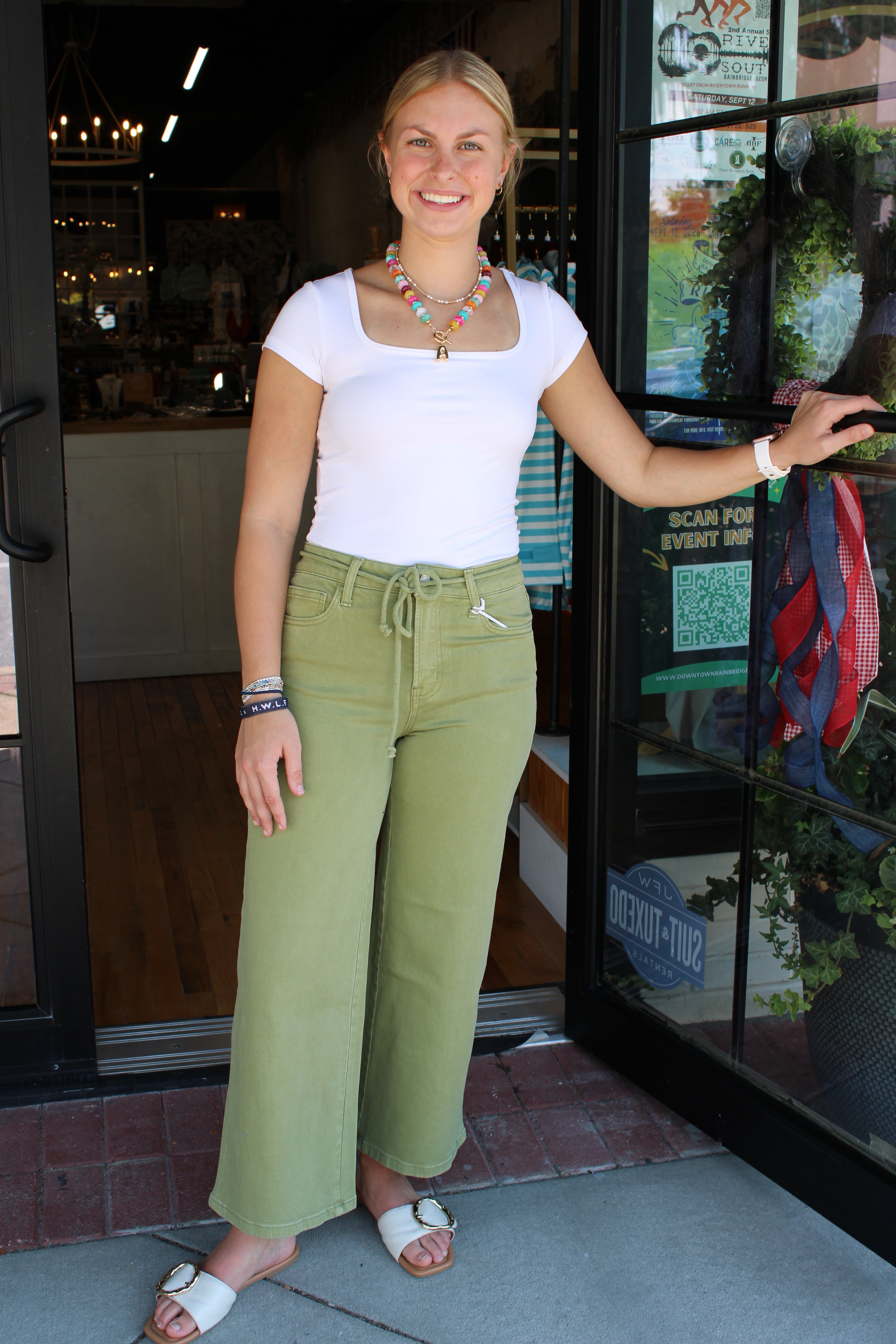 A woman stands in a storefront doorway wearing a white short-sleeve top and olive green wide-leg drawstring pants with white slide sandals.