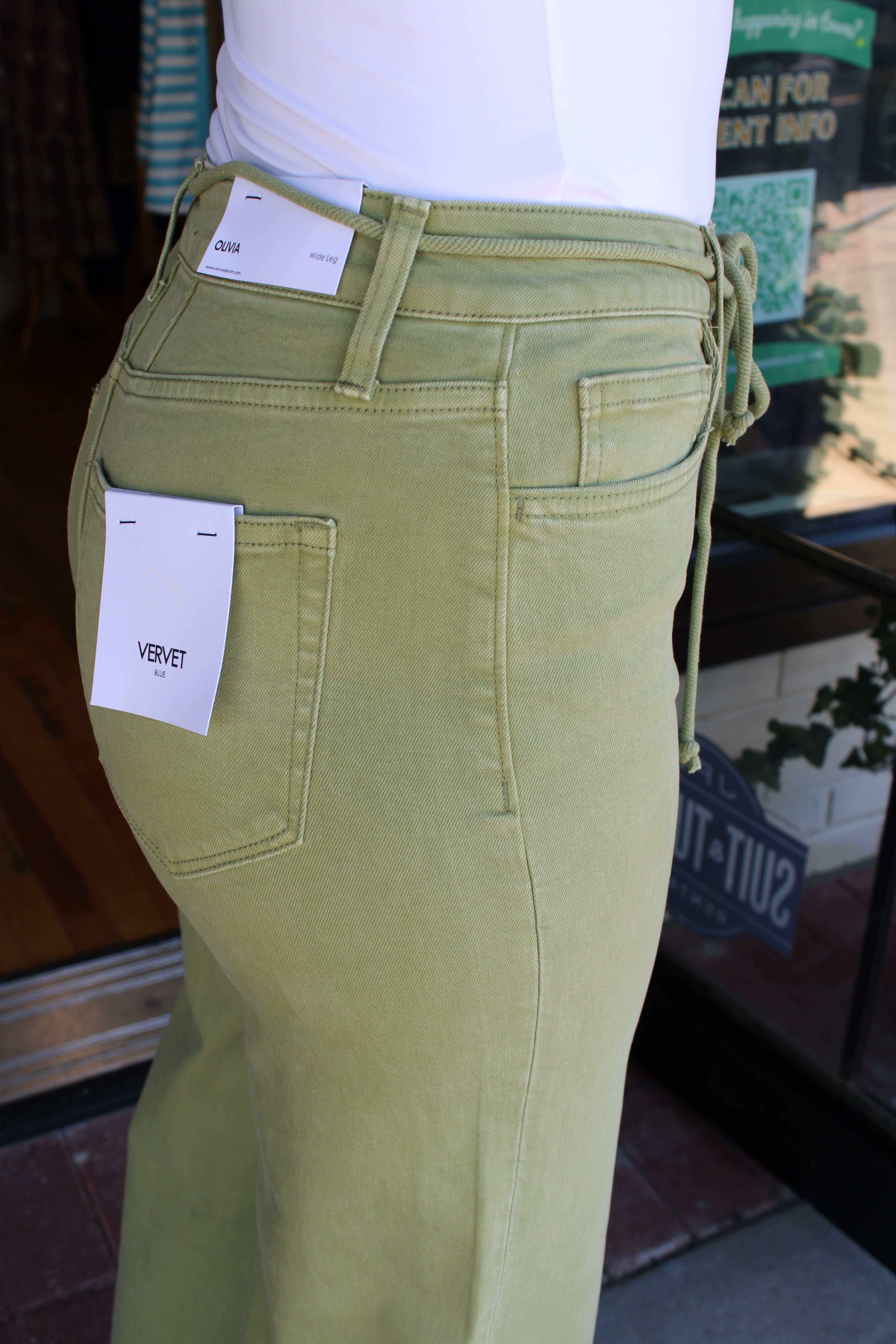 A close-up of a person wearing light olive green velvet drawstring jeans, showing the back pocket with a white brand tag that reads 'VERVET', a white top, and a storefront background.