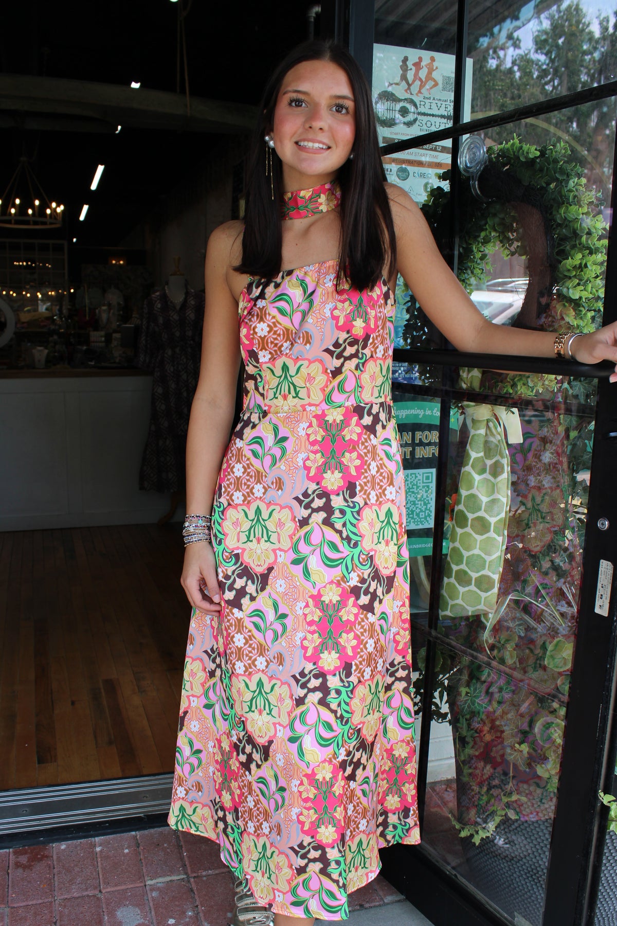 A woman wearing a colorful floral strapless maxi dress stands in the doorway of a shop, holding onto the black metal frame of the glass door.