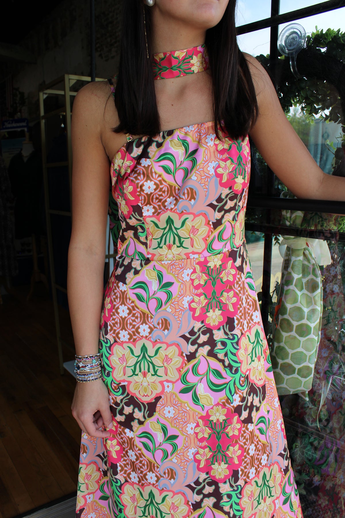 A woman wearing a sleeveless halter-neck dress with a vibrant retro floral pattern in pink, green, yellow, and brown on a light pink base, standing in a shop.