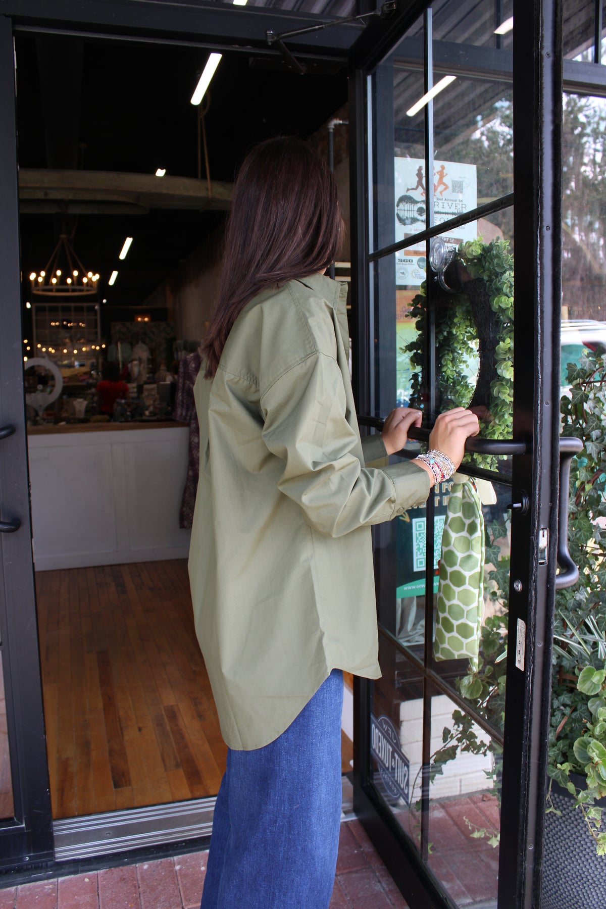 A woman wearing a light olive green button-up shirt and a blue skirt stands at an open glass storefront door, holding the handle. The background shows a cozy shop interior with wooden floors and warm lighting.