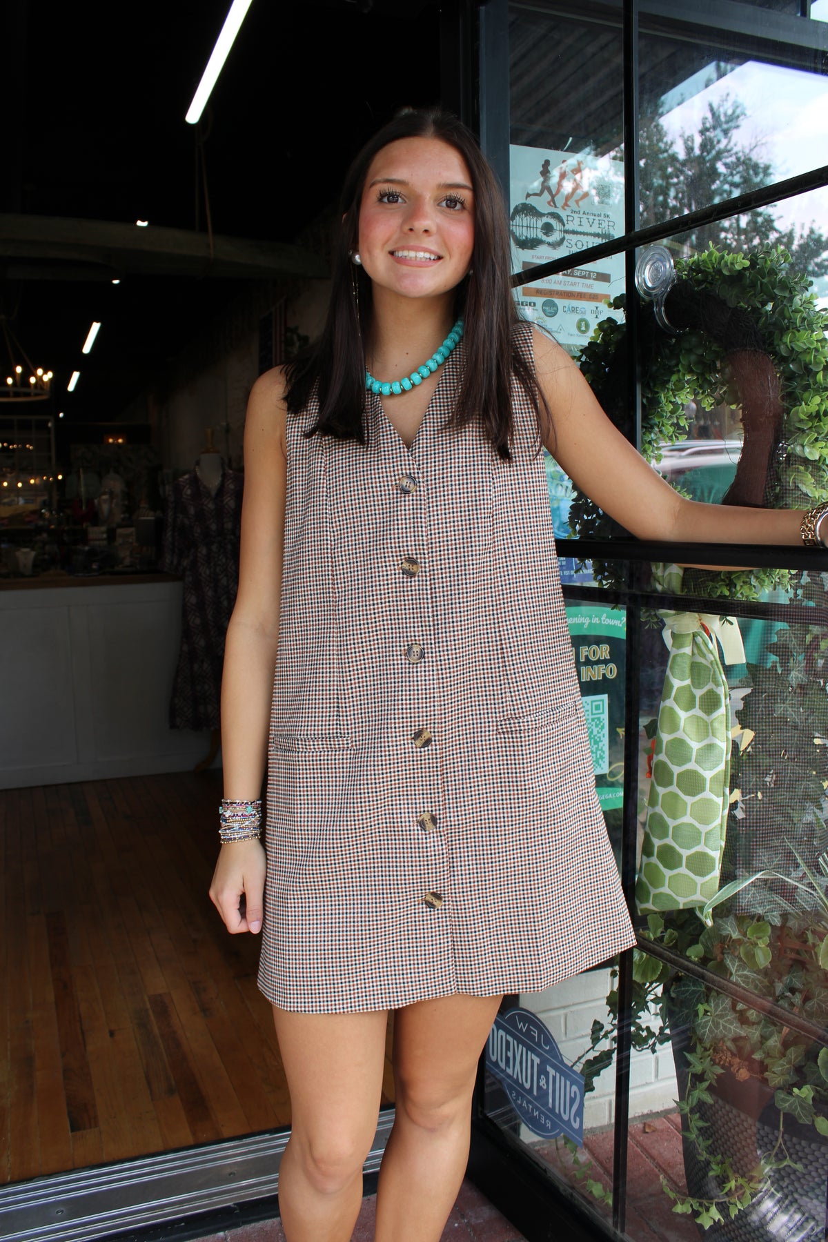 A woman wearing a sleeveless brown and beige checkered mini dress with front buttons, standing in front of a glass storefront window.