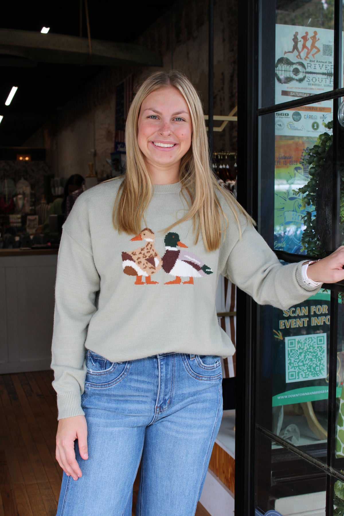 A smiling blonde woman wearing a sage green crewneck sweater with a colorful embroidered duck design on the front, paired with blue jeans, standing in a shop doorway.