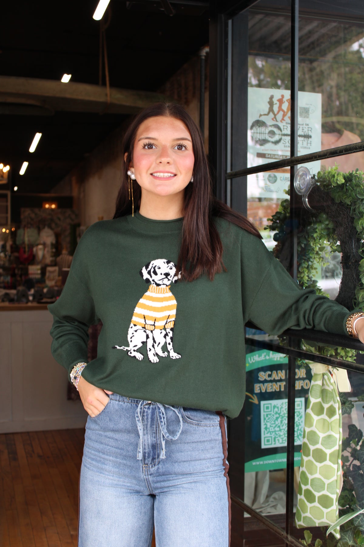 A woman wearing a dark green crewneck sweatshirt with a Dalmatian graphic wearing a yellow and white striped sweater, paired with light blue jeans, standing in a shop setting.