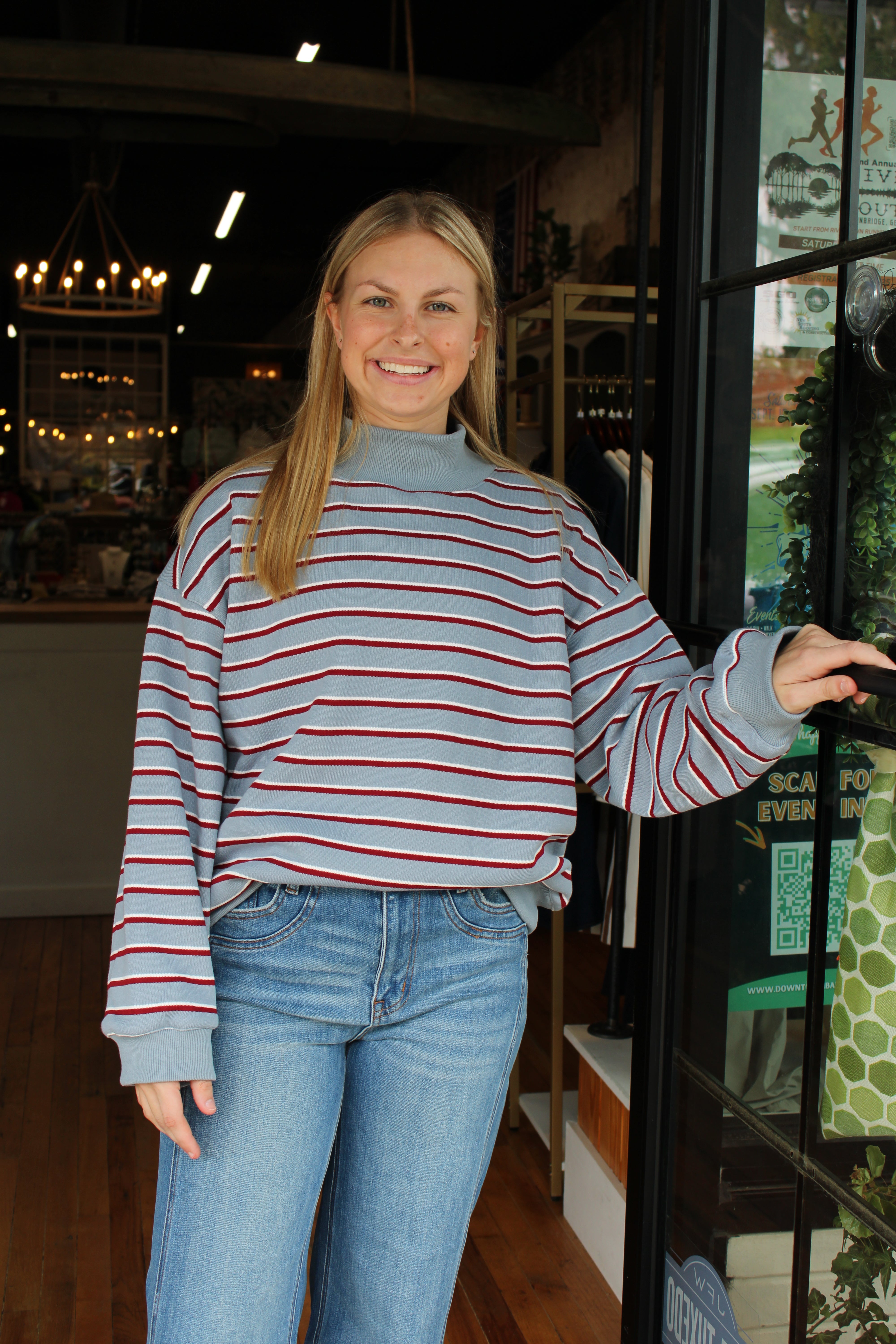 A woman with blonde hair stands in a shop doorway wearing a light blue mock-neck long-sleeve sweatshirt with thin red and navy horizontal stripes, paired with blue jeans.