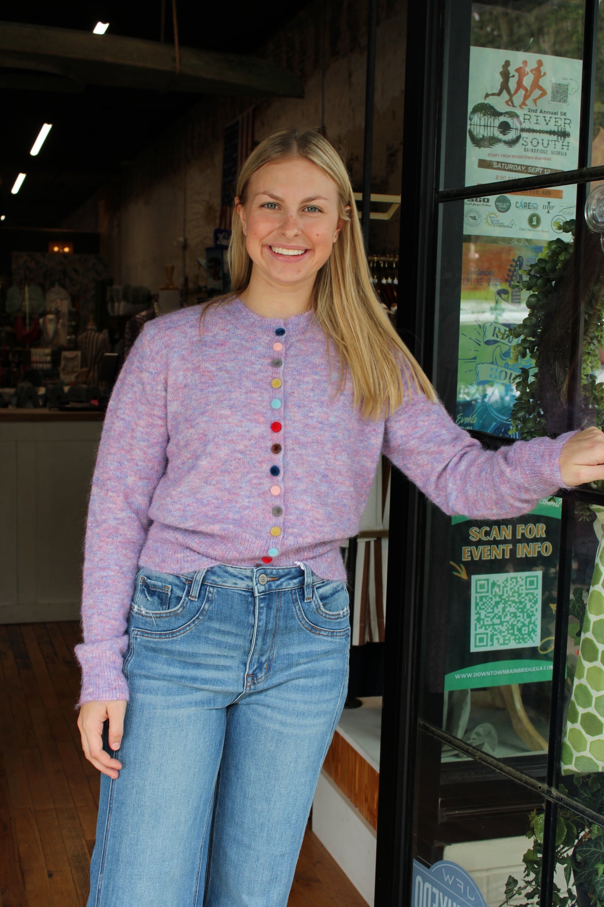 A smiling blonde woman wearing a lavender marled knit sweater with colorful buttons and blue jeans, standing in a shop doorway.