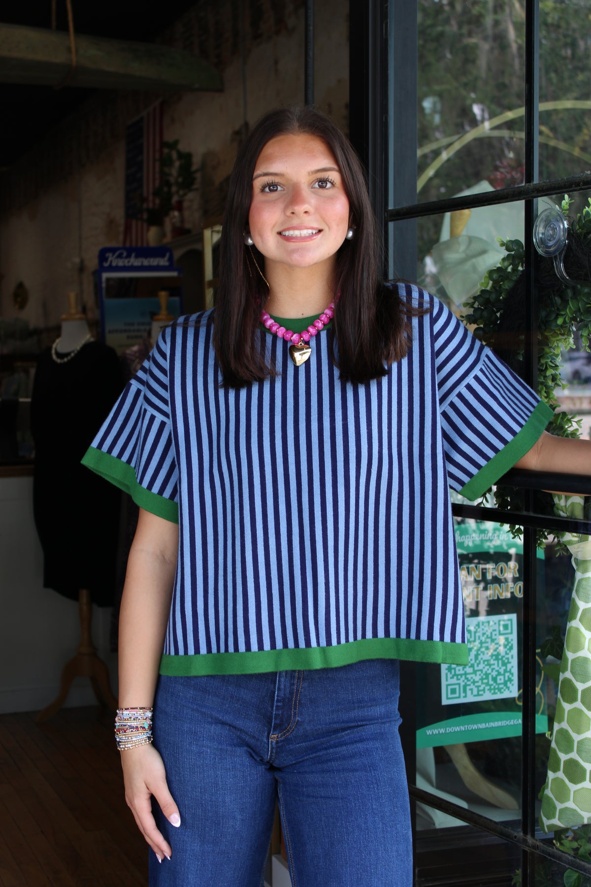A young woman with dark hair stands in a boutique doorway wearing a blue and white vertically striped short-sleeve top with green trim, paired with blue jeans.