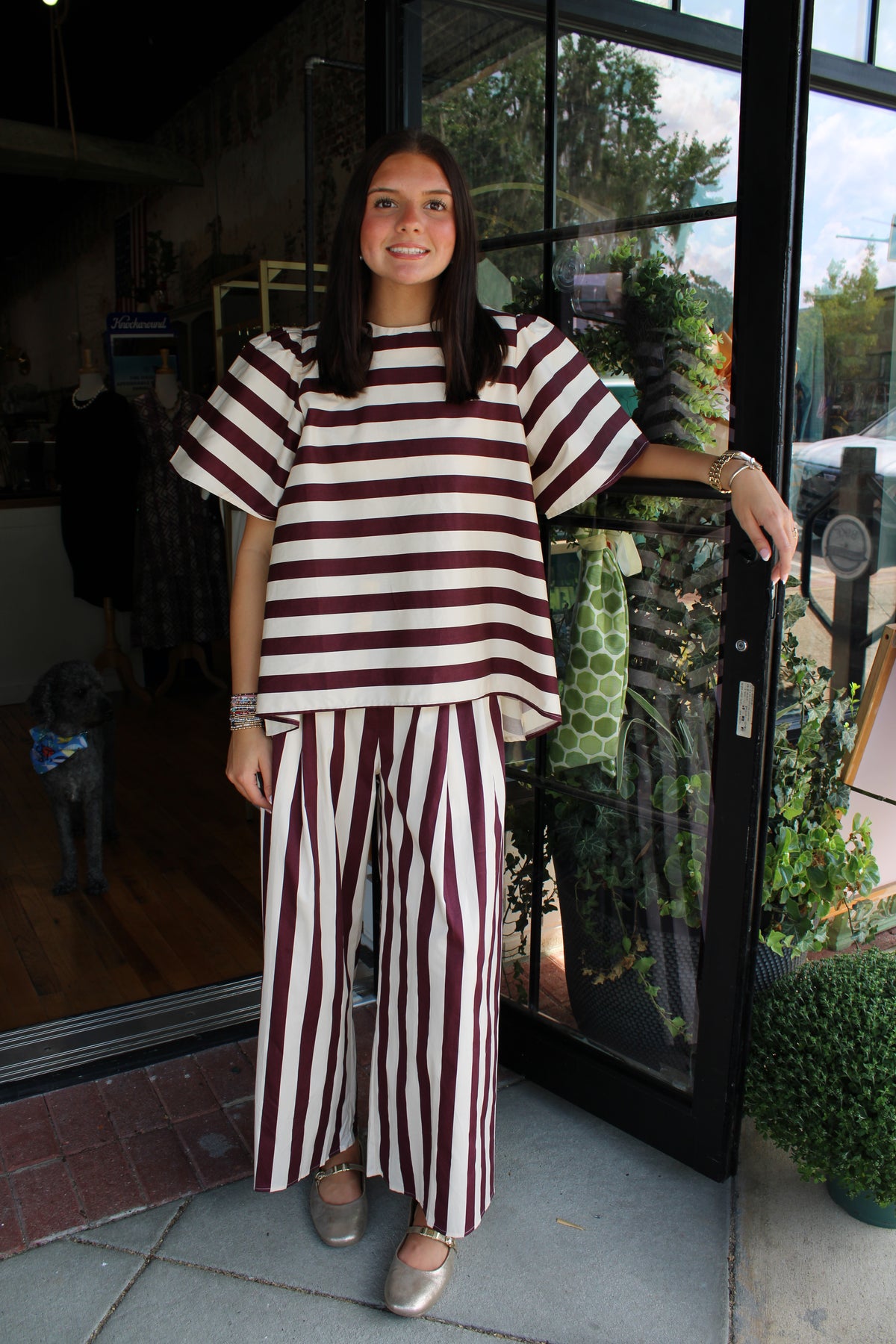 A woman stands on a storefront sidewalk wearing a matching two-piece outfit with horizontal burgundy and cream stripes on the top and vertical burgundy and cream stripes on the wide-leg pants.