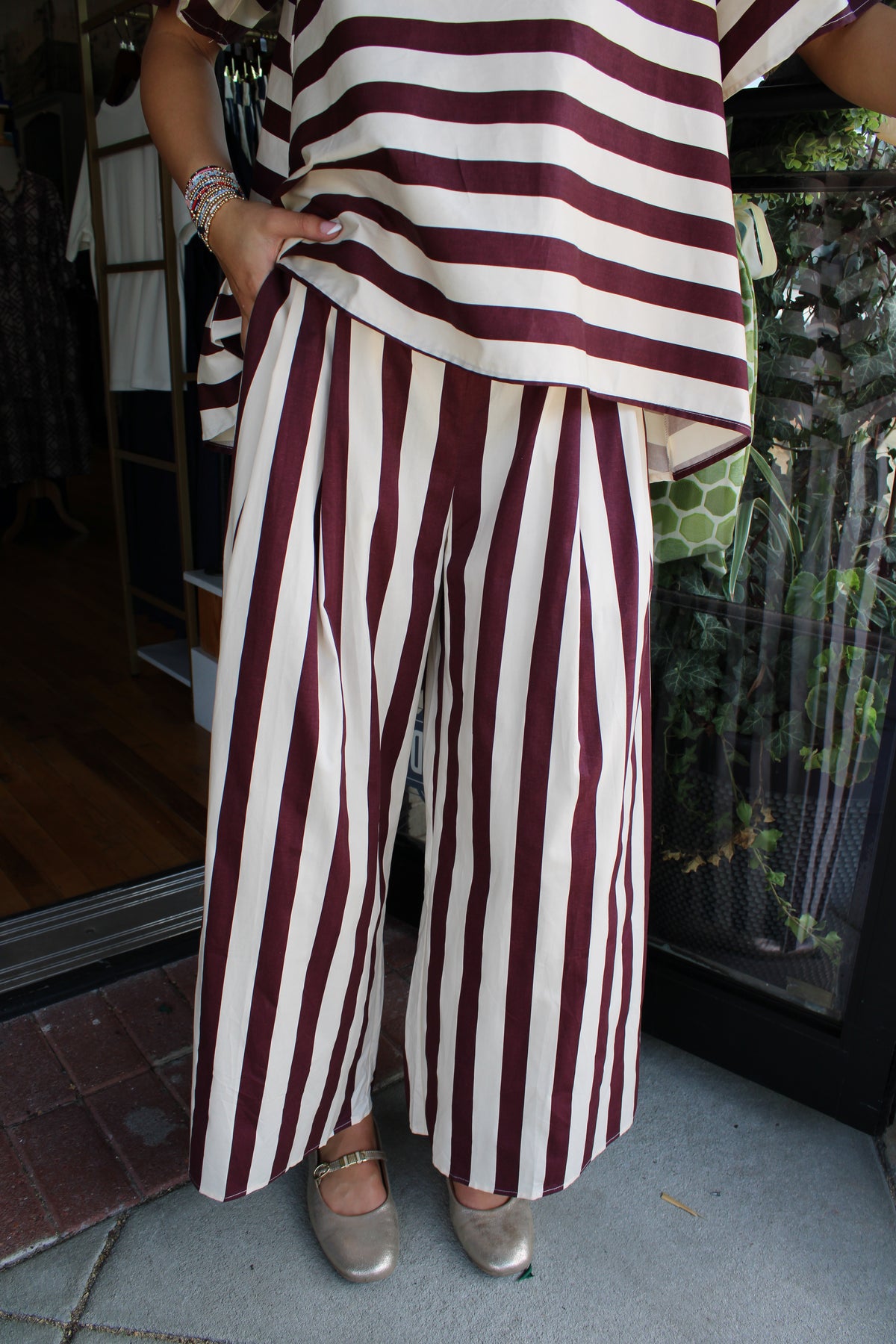A person wearing a matching burgundy and cream striped top and wide-leg pants, paired with metallic silver shoes, standing on a tiled floor in front of a glass door.